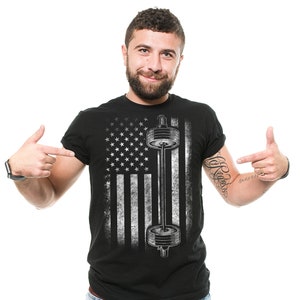 Puede incluir: Camiseta negra con un diseño desgastado de la bandera estadounidense y un gráfico de barra con pesas en blanco. La camiseta tiene cuello redondo y mangas cortas. El diseño está centrado en la parte delantera.