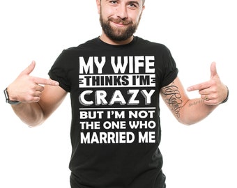 以下が含まれることがあります： 「MY WIFE THINKS I'M CRAZY BUT I'M NOT THE ONE WHO MARRIED ME」と白い文字で書かれた黒いTシャツ。人がTシャツを指しています。