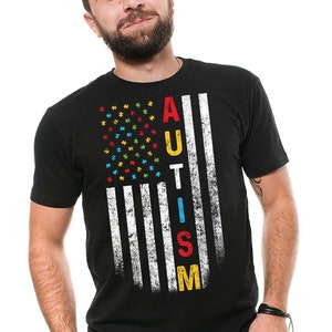 Puede incluir: Una camiseta negra con un dise&ntilde;o de bandera estadounidense desgastado. La bandera est&aacute; formada por piezas de rompecabezas en los colores de la cinta de concienciaci&oacute;n sobre el autismo: azul, rojo, amarillo y verde. La palabra "AUTISMO" est&aacute; escrita verticalmente en la bandera con letras blancas.