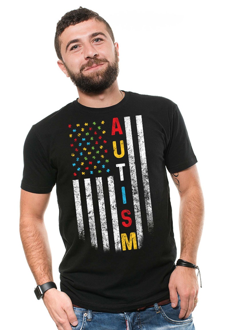 Puede incluir: Una camiseta negra con un dise&ntilde;o de bandera estadounidense desgastado. La bandera est&aacute; formada por piezas de rompecabezas en los colores de la cinta de concienciaci&oacute;n sobre el autismo: azul, rojo, amarillo y verde. La palabra "AUTISMO" est&aacute; escrita verticalmente en la bandera con letras blancas.