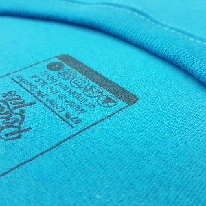 Puede incluir: Un primer plano de una etiqueta de camiseta azul con texto negro que dice "Hecho en los EE. UU. 97% algod&oacute;n 3% spandex de tela importada".
