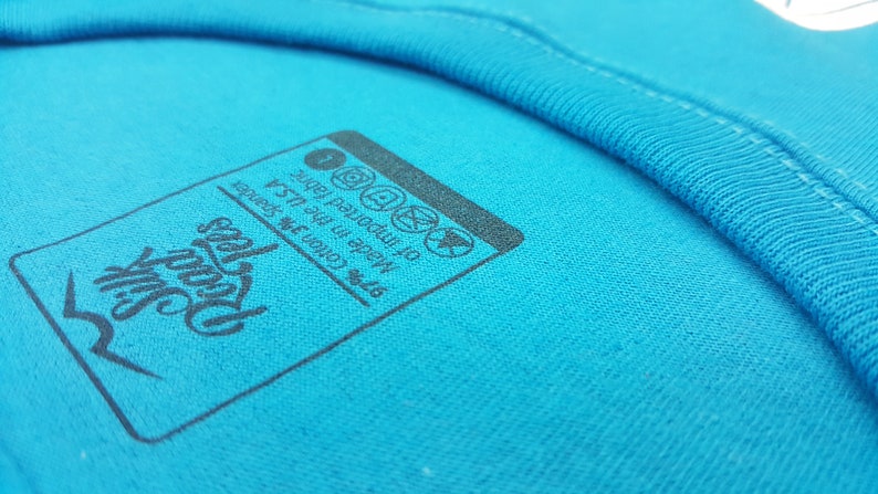 Puede incluir: Un primer plano de una etiqueta de camiseta azul con texto negro que dice "Hecho en los EE. UU. 97% algod&oacute;n 3% spandex de tela importada".