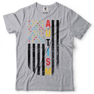 Puede incluir: Camiseta gris con un dise&ntilde;o de bandera estadounidense desgastado. La bandera est&aacute; compuesta por piezas de rompecabezas en azul, rojo, amarillo y verde. La palabra "AUTISMO" est&aacute; escrita verticalmente en la bandera en negro.
