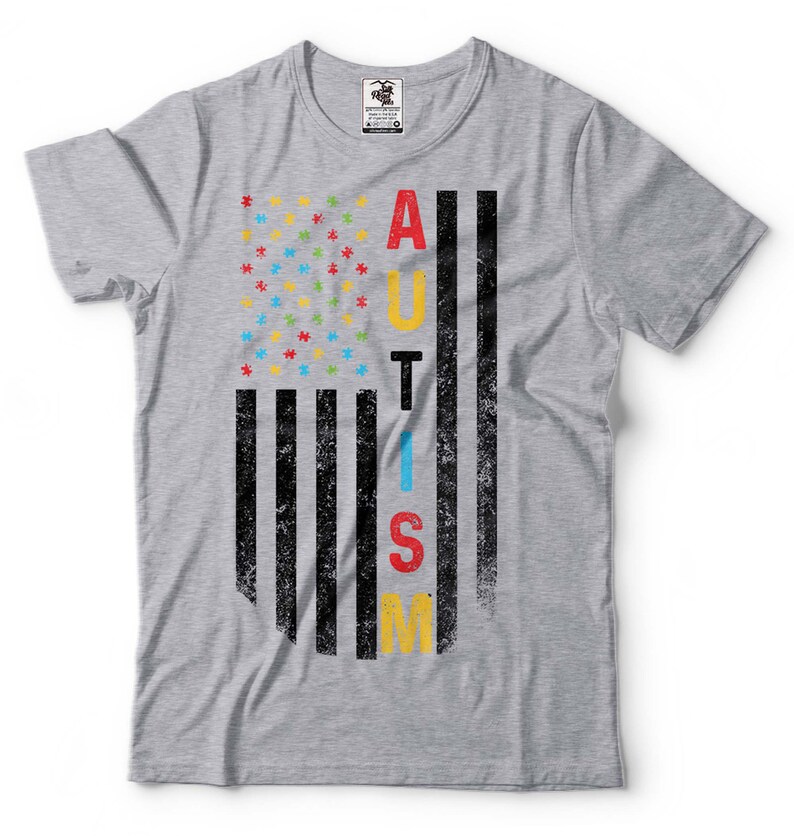 Puede incluir: Camiseta gris con un dise&ntilde;o de bandera estadounidense desgastado. La bandera est&aacute; compuesta por piezas de rompecabezas en azul, rojo, amarillo y verde. La palabra "AUTISMO" est&aacute; escrita verticalmente en la bandera en negro.