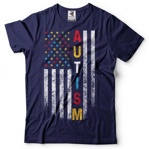 Puede incluir: Camiseta azul marino con un dise&ntilde;o de bandera estadounidense desgastado. La bandera est&aacute; formada por piezas de rompecabezas en los colores de la cinta de concienciaci&oacute;n sobre el autismo. La palabra "AUTISMO" est&aacute; escrita verticalmente en la bandera.