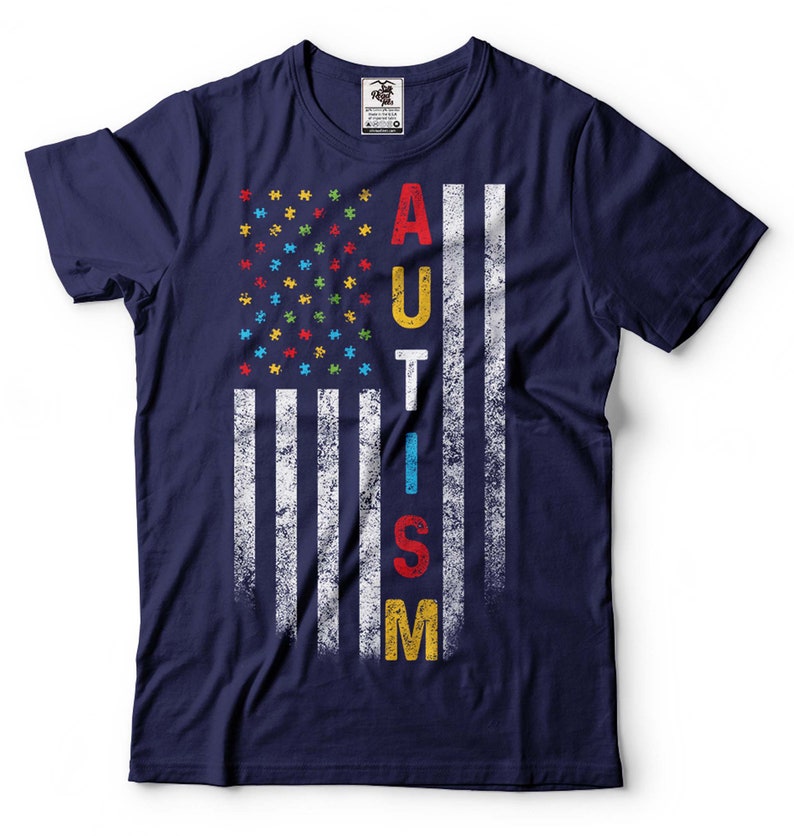 Puede incluir: Camiseta azul marino con un dise&ntilde;o de bandera estadounidense desgastado. La bandera est&aacute; formada por piezas de rompecabezas en los colores de la cinta de concienciaci&oacute;n sobre el autismo. La palabra "AUTISMO" est&aacute; escrita verticalmente en la bandera.