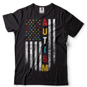Puede incluir: Camiseta negra con un dise&ntilde;o de bandera estadounidense desgastado. La bandera est&aacute; formada por piezas de rompecabezas en los colores de la cinta de concienciaci&oacute;n sobre el autismo. La palabra "AUTISMO" est&aacute; escrita verticalmente en la bandera con letras de colores.