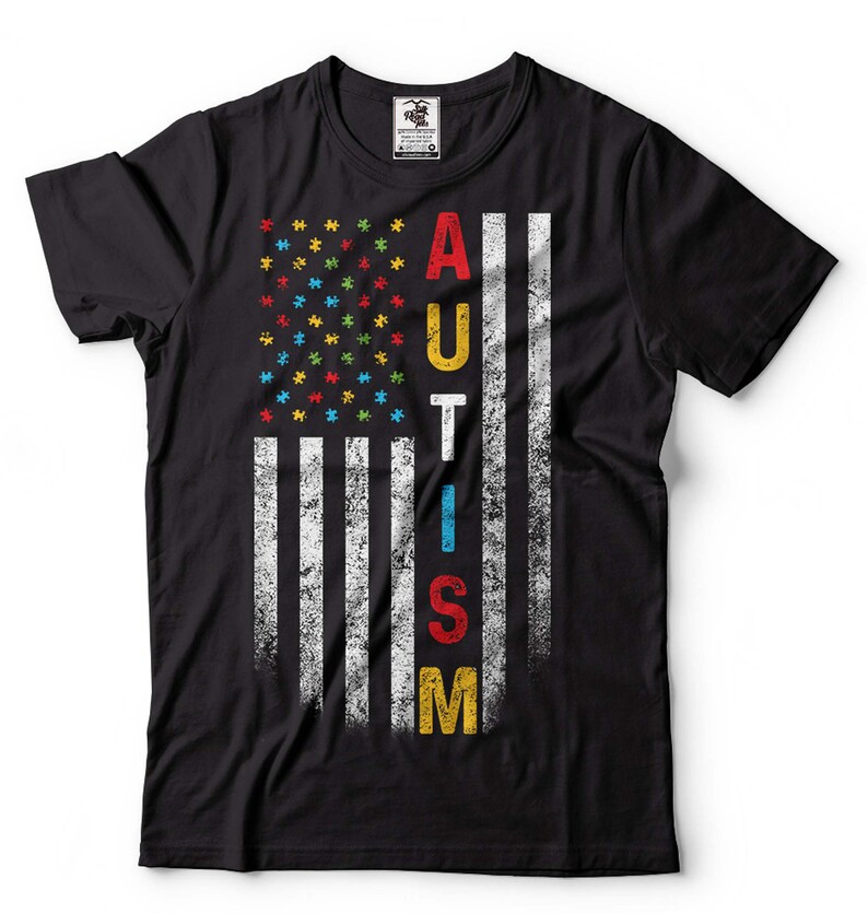 Puede incluir: Camiseta negra con un dise&ntilde;o de bandera estadounidense desgastado. La bandera est&aacute; formada por piezas de rompecabezas en los colores de la cinta de concienciaci&oacute;n sobre el autismo. La palabra "AUTISMO" est&aacute; escrita verticalmente en la bandera con letras de colores.
