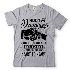 Puede incluir: Camiseta gris con un diseño gráfico negro que dice "Daddy & Daughters Not Always Eye To Eye But Always Heart To Heart". El gráfico también incluye dos puños chocando.