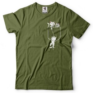 May include: A green t-shirt with a white graphic of three cats hanging from a pocket. The cats are drawn in a cartoon style and are looking up at the viewer. The text "I'm in the pocket" is printed on the shirt.