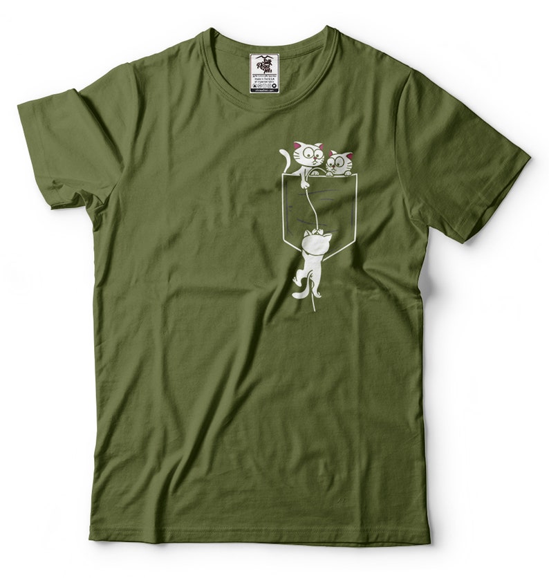 May include: A green t-shirt with a white graphic of three cats hanging from a pocket. The cats are drawn in a cartoon style and are looking up at the viewer. The text "I'm in the pocket" is printed on the shirt.