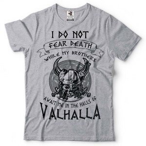 Puede incluir: Camiseta gris con un dise&ntilde;o gr&aacute;fico en blanco y negro de un casco vikingo con cuernos y el texto "No temo la muerte mientras mis hermanos me esperan en las salas de Valhalla".