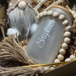 May include: A gift box filled with a gnome, a straw broom, a glass jar with a wooden lid, and a gold straw. The jar is frosted and has the name "Stephanie" written on it. The jar is surrounded by wooden beads.