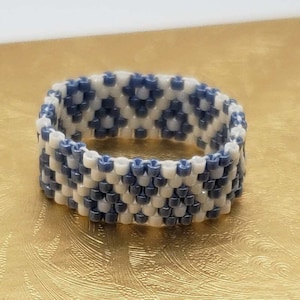 May include: A white and blue beaded ring with a geometric pattern. The ring is made of small, round beads that are woven together to create a unique design.