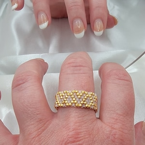 May include: A gold and white beaded ring with a woven pattern. The ring is worn on a person's finger.