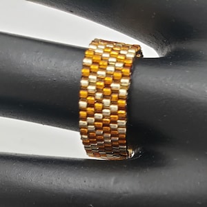 May include: A gold and brown beaded ring, woven in a herringbone pattern. The ring is on a black mannequin hand.