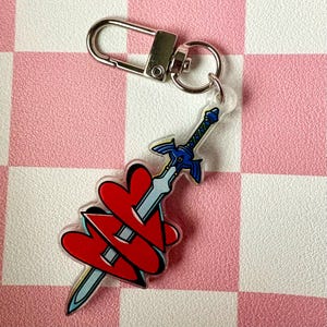 May include: A clear acrylic keychain featuring a stylized sword and three red hearts. The sword has a blue hilt and silver blade. The keychain includes a silver clasp and ring for attachment.