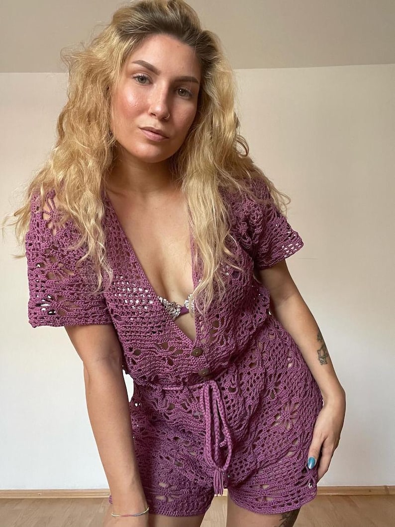 Op de afbeelding: Een mauve gebreide jumpsuit met een diepe V-hals, korte mouwen en een strik in de taille.
