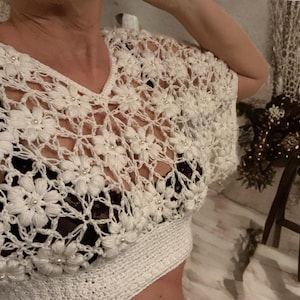 Peut inclure: Haut en crochet blanc avec un col en V. Le haut présente un motif floral avec de petits accents de perles. Le design en tissage ouvert est visible. L'ourlet inférieur est une bande blanche unie.