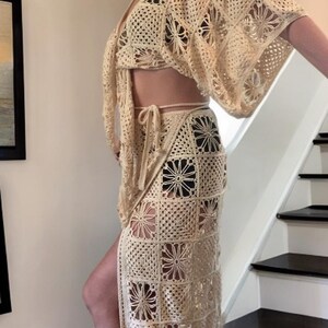 以下が含まれることがあります： クリーム色のクロシェ編みセット。ワイドスリーブのクロップトップ、ハイウエストのスリット入りラップスカート、お揃いのリボンが特徴です。四角いクロシェ編みパネルに花柄のデザインが施されています。