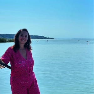 Op de afbeelding: Een vrouw in een roze gehaakte jurk staat aan een meer. Het meer is kalm en de lucht is blauw.