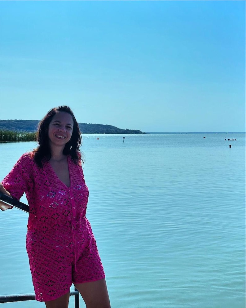 Op de afbeelding: Een vrouw in een roze gehaakte jurk staat aan een meer. Het meer is kalm en de lucht is blauw.