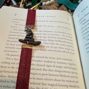 May include: A close-up of a book page with a red bookmark. The text on the page discusses the founding of Hogwarts School of Witchcraft and Wizardry. The bookmark features a bronze-colored Sorting Hat.