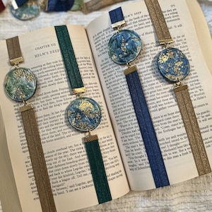 May include: Four handmade bookmarks with a blue, green, and gold color scheme. Each bookmark features a round, textured, painted design with gold accents. The bookmarks are attached to a ribbon with a tassel.