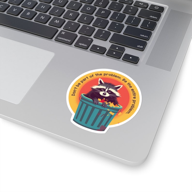 May include: A round sticker featuring a raccoon in a teal trash can, with the text "Don't be part of the problem. Be the entire problem." The raccoon is black and white with a mischievous expression. The sticker is on a laptop.