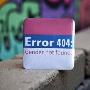 May include: A square badge with a pink top, blue stripe, and white base. The text reads "Error 404: Gender not found." The background shows colourful graffiti.