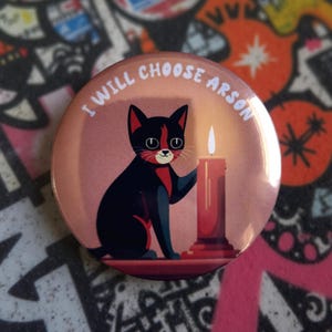 Può includere: Un bottone rotondo con un design a cartone animato di gatto e candela. Il gatto nero ha accenti rossi ed è seduto accanto a una candela rossa. Il testo "I WILL CHOOSE ARSON" è stampato attorno al bordo del bottone.