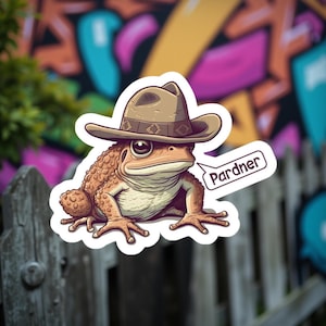 May include: A sticker of a cartoon frog wearing a brown cowboy hat. The frog has a textured, tan body and large eyes. A speech bubble says "Pardner". The background includes a wooden fence and colourful graffiti.