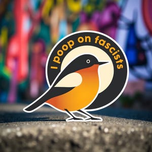Peut inclure: Un autocollant représentant un oiseau stylisé avec un plumage orange et noir. L'oiseau est placé sur un cercle crème avec le texte "I poop on fascists" dans un anneau noir. Le fond est un mur de graffiti flou.