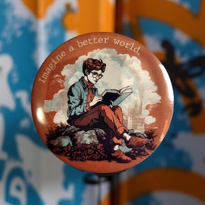Pode incluir: Botão redondo com uma ilustração vintage de uma pessoa lendo um livro, usando óculos e sentada em uma rocha. O botão tem o texto "Imagine a better world". O esquema de cores inclui tons de marrom, verde e azul.