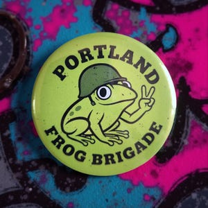 May include: A round, lime green button with the words "PORTLAND FROG BRIGADE" in black. The button features a cartoon frog wearing a green army helmet and giving a peace sign. The background is a mix of teal, pink, and black.