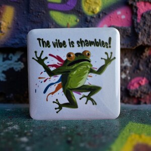 Peut inclure: Bouton carré blanc avec une illustration de grenouille verte et le texte "The vibe is shambles!". La grenouille a des yeux orange et est entourée d'éclaboussures de peinture colorées. Le bouton est sur une surface texturée.