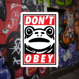 May include: A rectangular sticker with a black and white frog face design and the words "DON'T OBEY" in red and white. The sticker is set against a backdrop of a graffiti-covered wall.