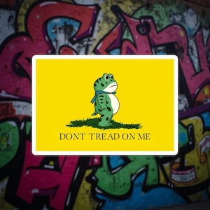 Può includere: Un adesivo giallo con un ranocchio dei cartoni animati con una sciarpa blu, in piedi sull'erba. Il testo "DONT TREAD ON ME" è sotto la rana. Lo sfondo è un muro di graffiti colorato.