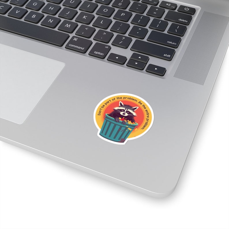 May include: A round sticker featuring a raccoon in a teal trash can, set against an orange and yellow background. The text reads, "Don't be part of the problem. Be the entire problem." The sticker is placed on a laptop.