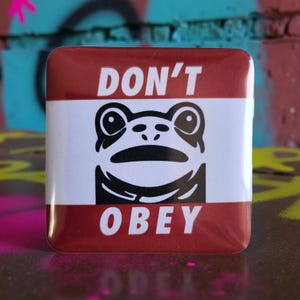 May include: Square button with a red border and the words "DON'T OBEY" in white. The center features a black and white graphic of a frog's face.