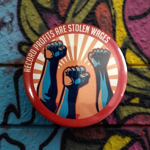Peut inclure: Un bouton rond rouge avec le texte "RECORD PROFITS ARE STOLEN WAGES". Le design présente trois poings levés en bleu et orange sur un fond rayonnant orange et blanc.