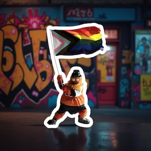 Peut inclure: Un autocollant représentant une mascotte en tenue orange et noire, tenant un drapeau avec un design arc-en-ciel et progressiste. Le fond montre du graffiti sur un mur.