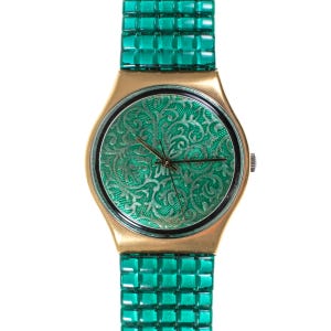Op de afbeelding: Een goudkleurig horloge met een groene wijzerplaat met een zilveren bloemenontwerp. Het horloge heeft een bijpassende groene band. De wijzerplaat heeft een diameter van ongeveer 3,2 cm.