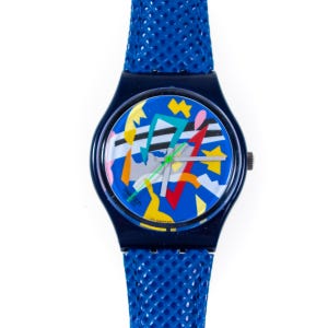 Può includere: Un orologio da polso Swatch con cassa in plastica blu scuro e cinturino blu testurizzato. Il quadrante presenta un vivace design grafico astratto con forme gialle, rosse, azzurre e nere su sfondo blu, con lancette argentate e una lancetta verde.
