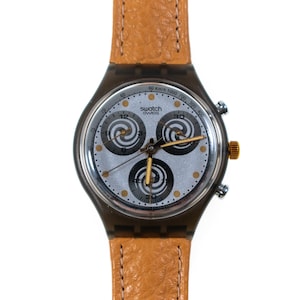 Op de afbeelding: Een Swatch Swiss chronograaf horloge met een lichtbruine leren band. De wijzerplaat is zilverkleurig met drie subwijzerplaten en gouden accenten. Het horloge heeft een zwarte kunststof kast en een goudkleurige kroon.