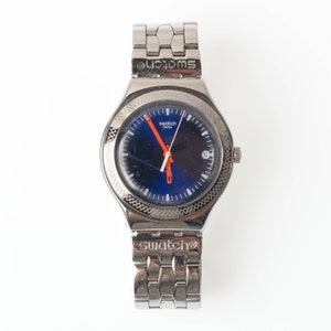 May include: A silver Swatch watch with a blue face and orange hands. The watch has a silver metal band.