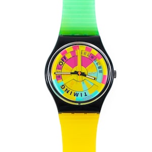 Può includere: Un orologio da polso Swatch con cassa nera, cinturino superiore verde traslucido e cinturino inferiore giallo a tinta unita. Il quadrante presenta un motivo segmentato colorato in rosa, giallo e blu con il testo OFF e TIMING stampato.