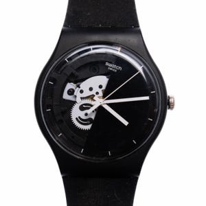 May include: A black Swatch watch with a white face and a partially exposed mechanical movement. The watch has a black rubber strap.