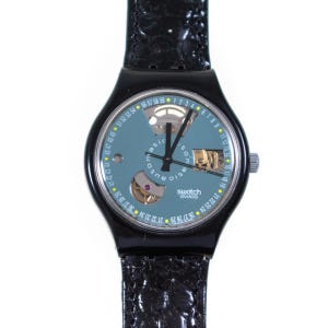 Puede incluir: Un reloj de pulsera negro con correa negra texturizada y caja redonda negra. La esfera es de color azul pizarra con una escala circular de números del 1 al 59, manecillas negras y dos aberturas que muestran piezas mecánicas. Swatch Swiss está impreso.