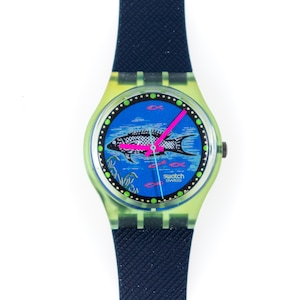 Può includere: Un orologio da polso con cinturino blu scuro testurizzato e cassa verde chiaro traslucida. Il quadrante blu presenta un'illustrazione di un pesce in bianco e nero, piccole icone di pesci rosa e lancette rosa acceso. La scritta Swatch è in bianco.